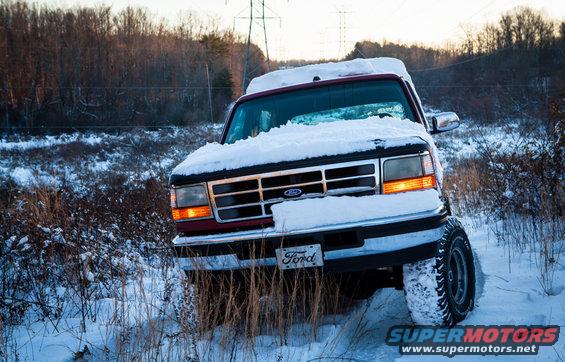 Red Ford truck covered in snow, front 3/4 view, with a visible Ford logo on the grille and a license plate