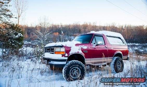 Red 1995 Ford Bronco covered in snow, parked in a snowy field with trees in the background