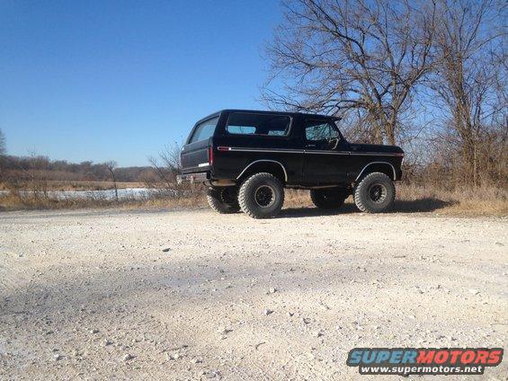 Black Ford Bronco parked on a dirt road, facing away from the camera. Bare trees and a frozen body of water are in the background