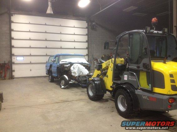 Blue pickup truck with snow on hood, front end disassembled, being towed by a yellow Wacker Neuson loader inside a garage