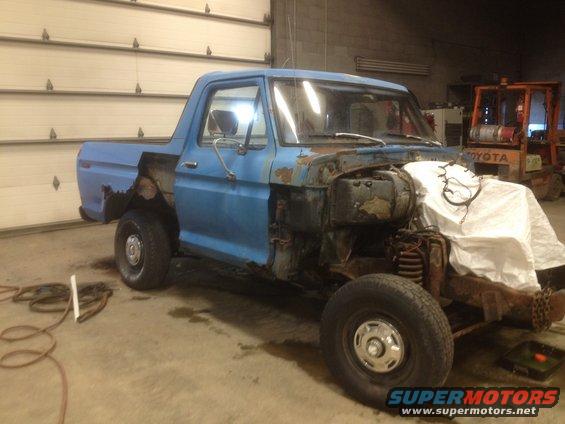 Blue pickup truck with front end disassembled, covered with a white sheet. A forklift is in the background
