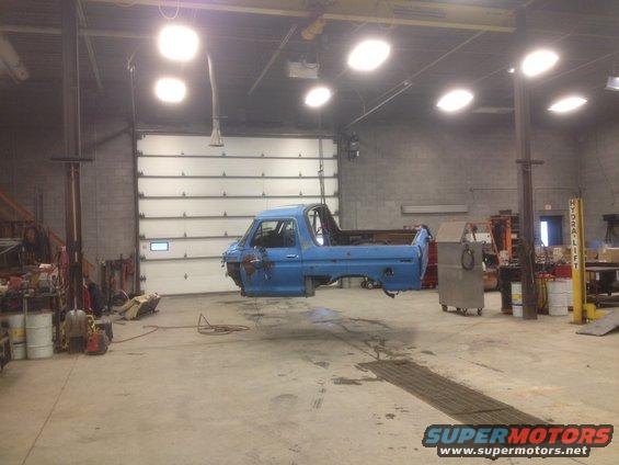 Blue pickup truck body, likely from the 1970s, suspended by a hoist in a garage