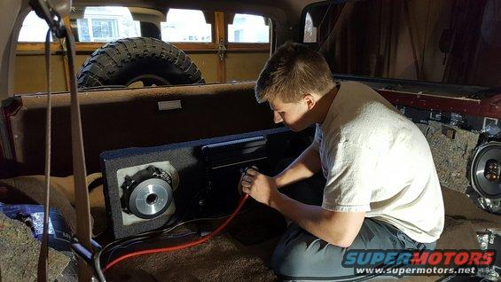 Person wiring a subwoofer and amplifier inside a vehicle's cargo area, spare tire visible