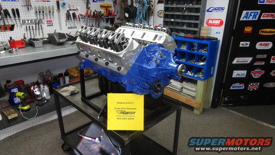 Blue V8 engine on a stand in a workshop, with a yellow sign