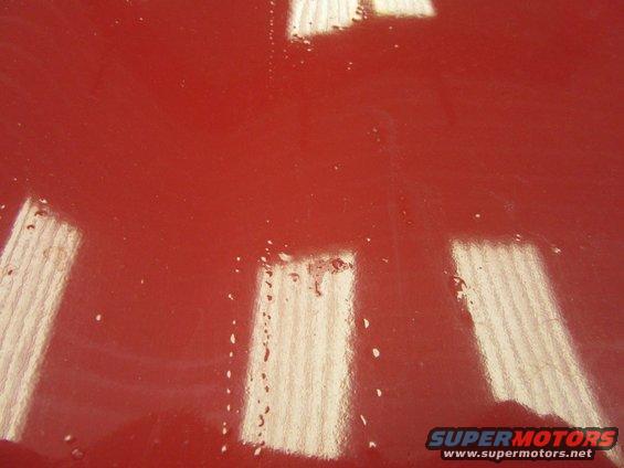 Close-up of a red car's hood with water droplets and reflections. Supermotors.net logo visible