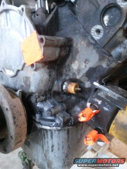 Engine block with a sensor and orange tape covering openings. Supermotors.net logo visible