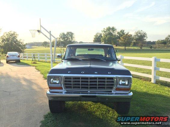 Front view of a dark blue Ford truck parked in a driveway with a basketball hoop behind it. Another car is visible in the background