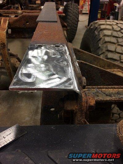 Close-up of a metal bracket being welded onto a rusty vehicle frame. A ruler is visible in the foreground