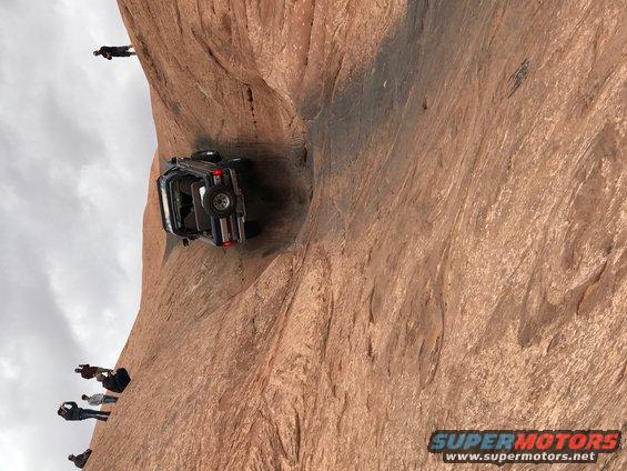 Dark blue Jeep Wrangler on its side on a steep rock face, with people watching.