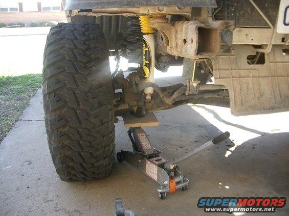 Front tire and suspension of a vehicle being lifted by a hydraulic jack. A wooden block is under the suspension