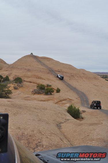 Several off-road vehicles driving up a steep, rocky trail on a desert landscape under a cloudy sky