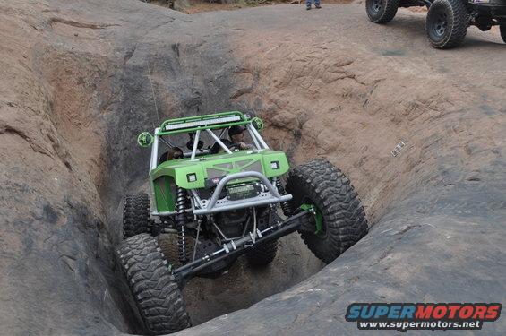 Green off-road buggy navigating a steep rock obstacle, with another vehicle visible in the background