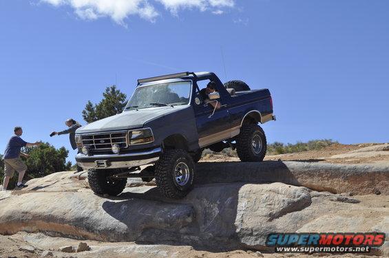 Ford Bronco climbing rocks on a trail, driver visible, two people watching