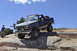 1991 Ford Bronco - A lifted Ford Bronco navigates a rocky obstacle on an off-road trail under a clear blue sky.