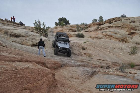 Silver Jeep Wrangler ascending a steep, rocky desert trail with people watching