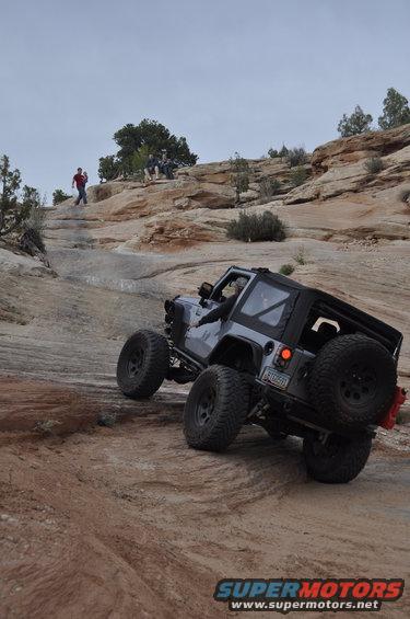Gray Jeep Wrangler with oversized tires climbing a rocky incline, spare tire mounted on the back