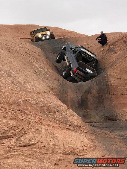 Dark blue SUV being towed up a rocky hill by a yellow Jeep