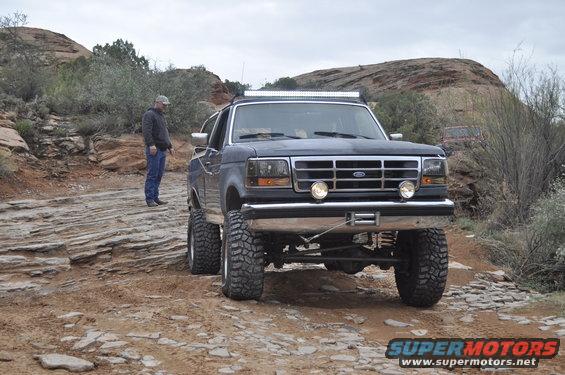 Ford F-Series truck on rocky off-road trail with a person standing nearby. Another vehicle is visible in the background