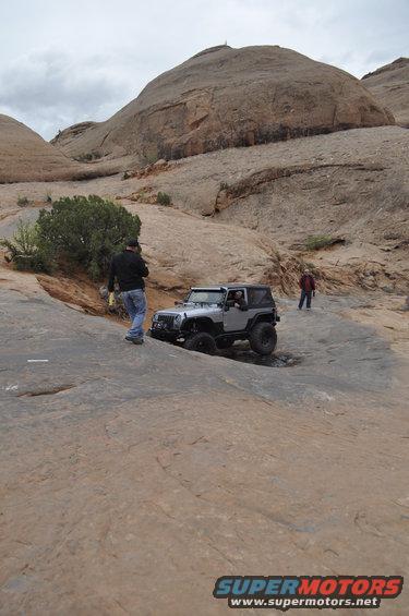 Silver Jeep Wrangler driving on rocky terrain with two people nearby
