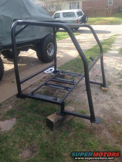 Black metal roll cage frame sitting on blocks next to a covered vehicle. A white SUV is parked in the background