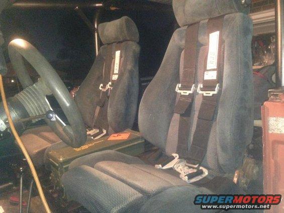 Interior of a vehicle with dark grey seats and racing harnesses, a green ammo box on the floor, and a large steering wheel