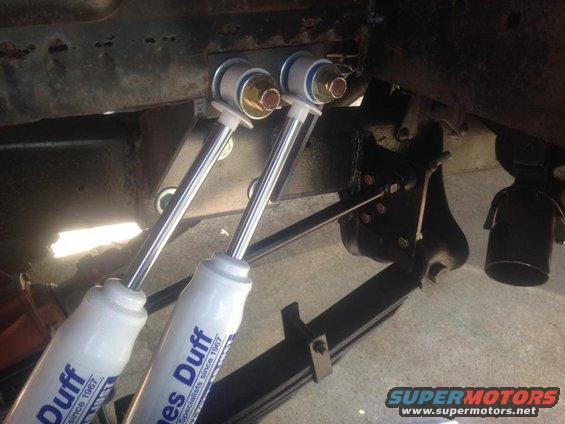 Close-up of two white shock absorbers with blue lettering, attached to a vehicle's rusty frame and leaf springs
