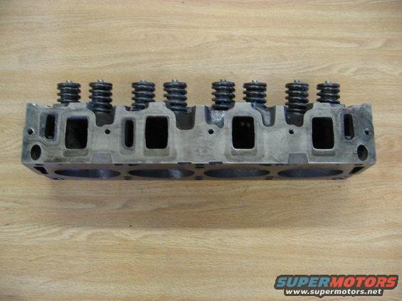 Engine cylinder head with valve springs on a wooden surface. Supermotors.net logo in the corner