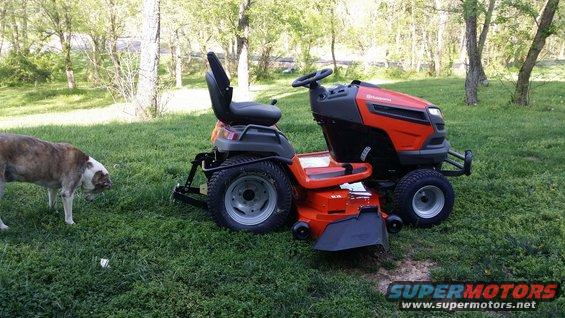 Orange and black Husqvarna riding mower parked on grass with a dog nearby