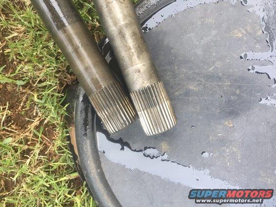 Two metal shafts with splined ends resting on a dark surface with water and grass in the background