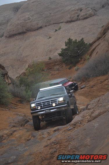 Dark Ford Bronco climbing rocky trail, another vehicle behind it