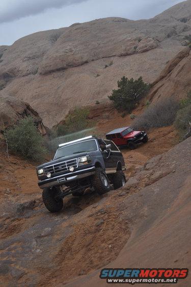Dark blue Ford Bronco climbing a rocky incline, front-left quarter view. Another red Jeep is behind it