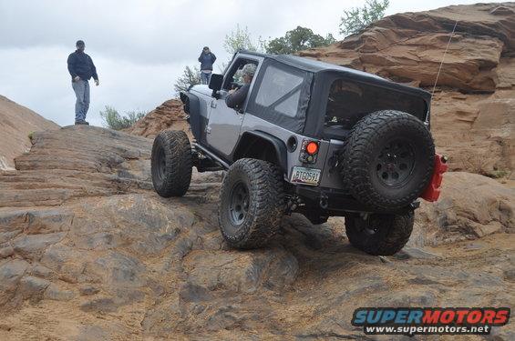 Silver Jeep Wrangler with a soft top navigating rocky terrain, rear view