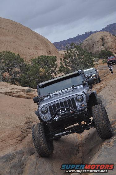 Jeep Wrangler with aftermarket lights and winch climbing rocks, other vehicles and people in background
