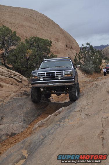 Dark blue Ford Bronco with light bar on roof climbing a rocky trail; another vehicle visible in background