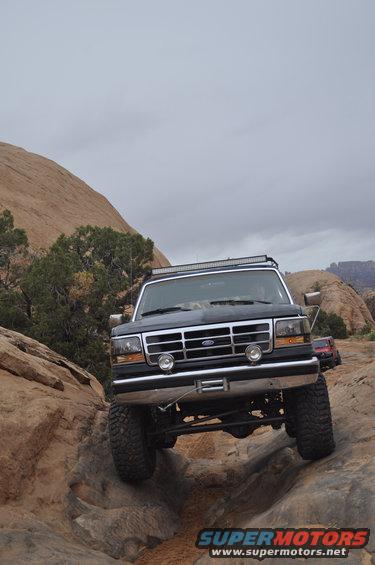 Dark blue Ford Bronco with a light bar on the roof, driving over rocks. Front grille and headlights are visible