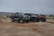 1991 Ford Bronco - Three modified off-road vehicles are parked in a dusty, arid landscape under an overcast sky.