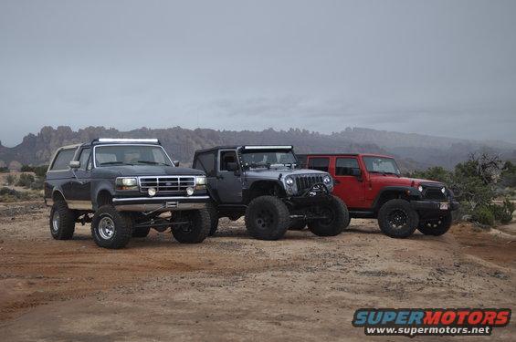 Dark blue Ford Bronco with a light bar and lifted suspension, parked on dirt with two Jeeps behind it