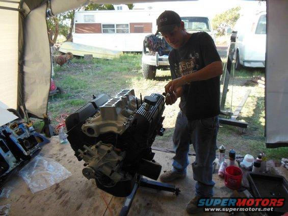 Man working on a V8 engine on an engine stand. Other engine parts and tools are visible