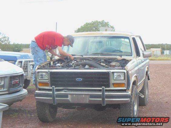 Blue Ford Bronco with hood open, mechanic working on engine. Front view