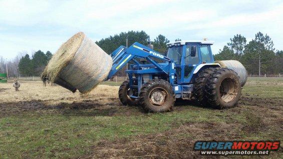 Blue Ford tractor lifting a large round hay bale with its front loader in a muddy field