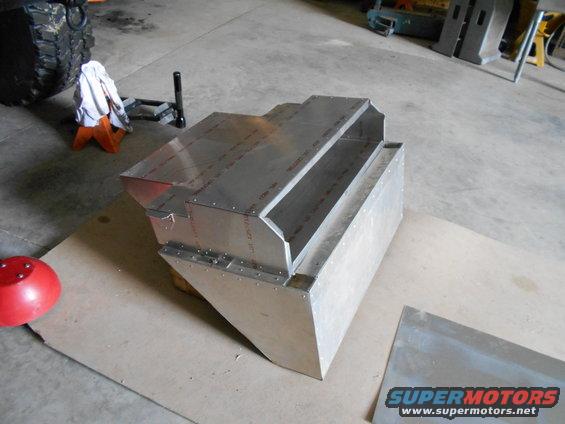 Aluminum fabricated part with visible rivets and seams, resting on cardboard in a workshop