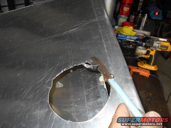 Tin snips cutting a circular hole in a large, scratched metal panel. Tools are visible in the background