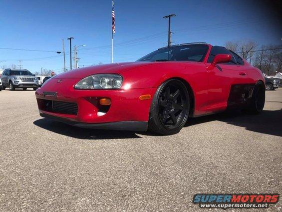 Red 1994 Toyota Supra parked on asphalt, front quarter view. Another SUV is visible in the background