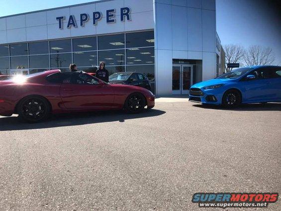Red sports car with black wheels parked in front of a dealership, next to a blue hatchback