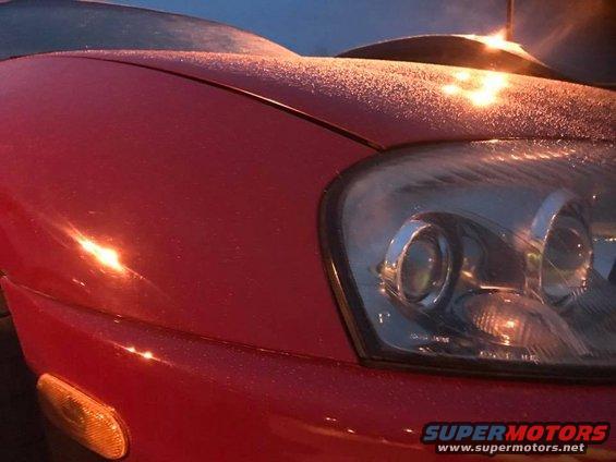 Red car hood and headlight with frost, showing a portion of the front bumper and turn signal