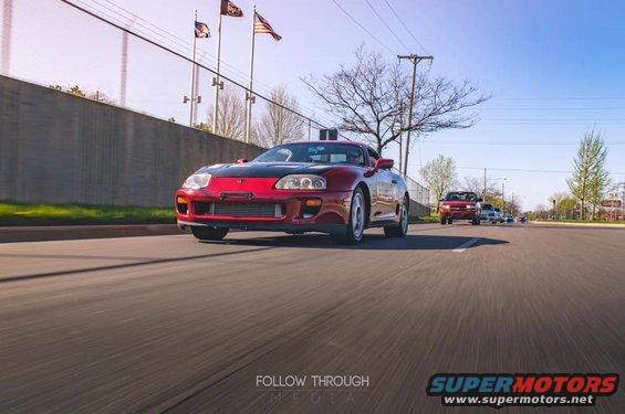 Red Toyota Supra with black hood driving on a road, front 3/4 view
