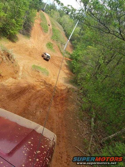 Red vehicle hood in foreground, white SUV climbing muddy hill in off-road scene