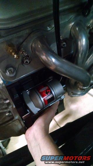 Hand installing a silver and red starter motor onto an engine block with chrome exhaust pipes