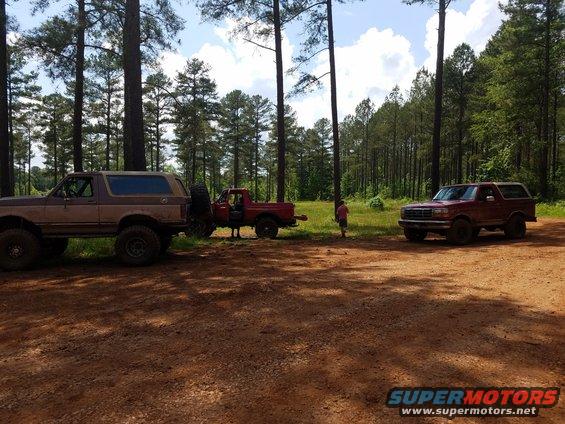 Brown 1987 Ford Bronco parked on dirt road with red pickup and red Bronco in background