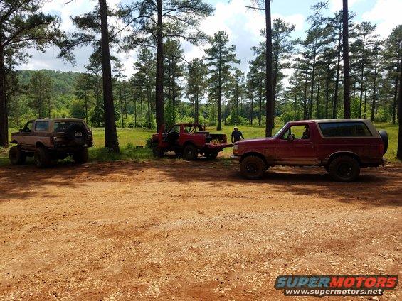 Three SUVs parked on a dirt road near trees. One is red, one is maroon, and one is tan
